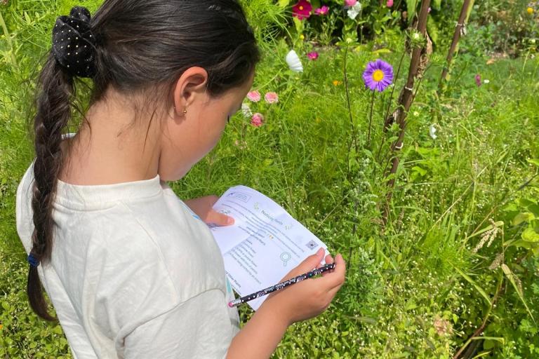 A child stands in a garden holding a nature activity sheet and pencil while observing plants and flowers.